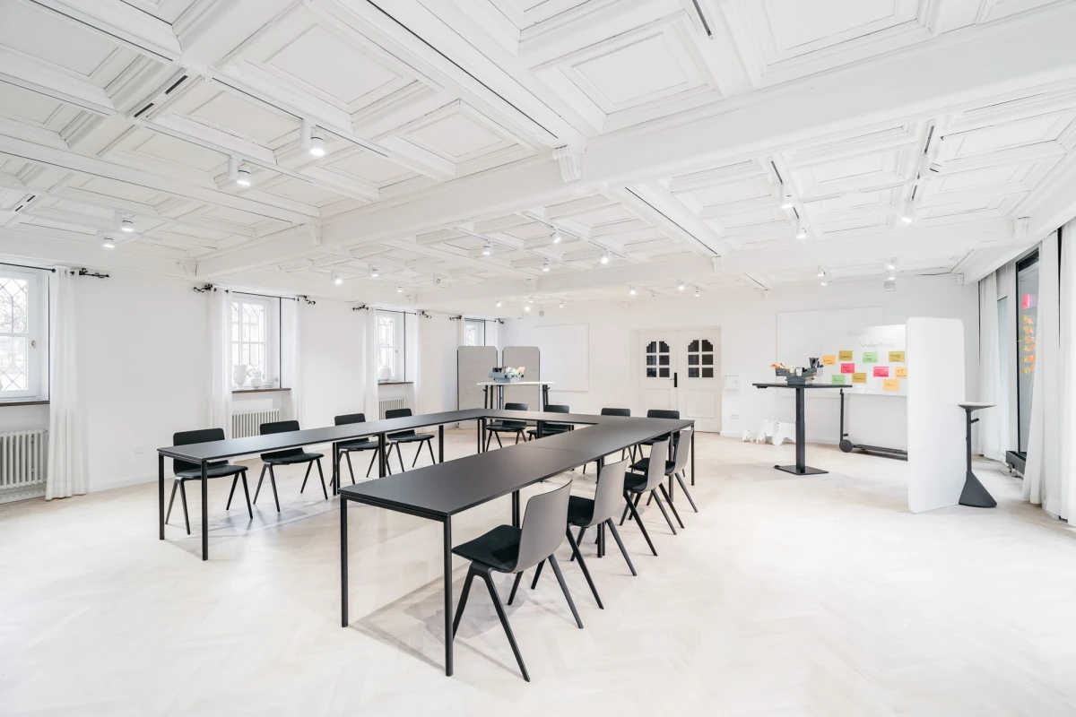 Heller, minimalistischer Seminarraum mit langen schwarzen Tischen und passenden St&uuml;hlen. Wei&szlig;e W&auml;nde und Decke mit Oberlicht, gro&szlig;e Fenster sorgen f&uuml;r nat&uuml;rliches Licht. An der Wand h&auml;ngen bukte Haftnotizen, ein Whiteboard steht bereit f&uuml;r Besprechungen und Workshops.