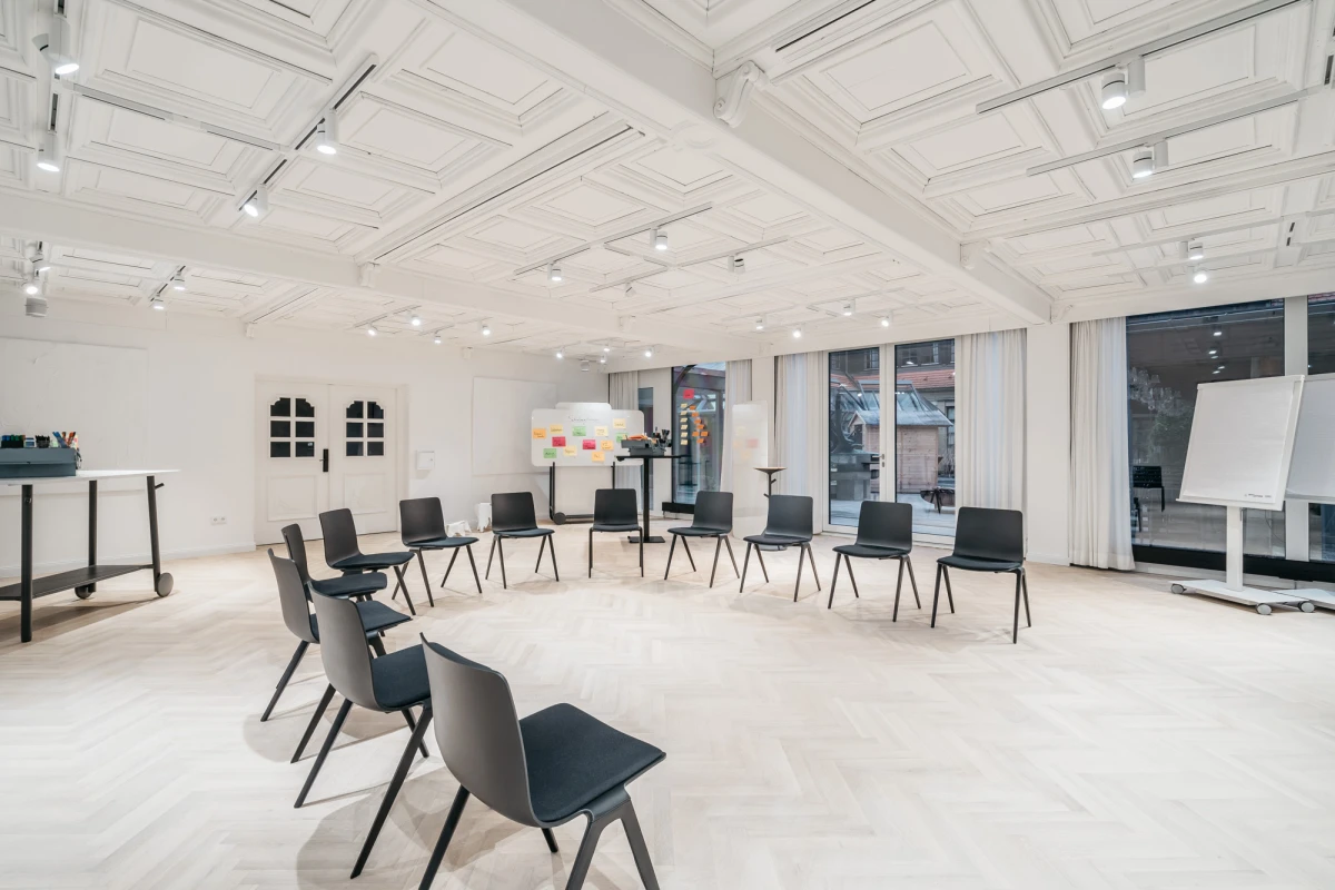Heller, minimalistischer Seminarraum mit wei&szlig;en W&auml;nden und Decke, ausgestattet mit schwarzen St&uuml;hlen, die in einem Halbkreis angeordnet sind. Gro&szlig;e Fenster lassen viel nat&uuml;rliches Licht herein, und Deckenspots sorgen f&uuml;r zus&auml;tzliche Beleuchtung. Ein Whiteboard an der Seite deutet auf einen Schulungs- oder Besprechungsraum hin.