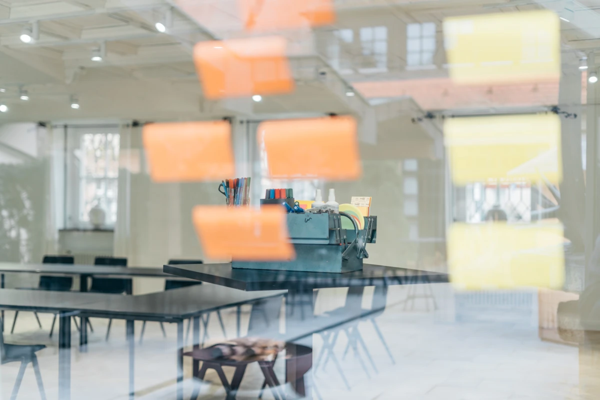 Blick in einen lichtdurchfluteten Besprechungsraum mit Glastisch und orangefarbenen Haftnotizen im Vordergrund. Moderne B&uuml;roeinrichtung mit St&uuml;hlen und ged&auml;mpftem Tageslicht, das durch gro&szlig;e Fenster f&auml;llt.