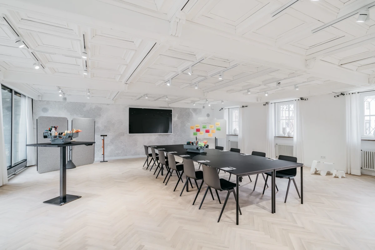 Moderner, heller Besprechungsraum mit gro&szlig;em schwarzem Tisch und schwarzen St&uuml;hlen. Wei&szlig;e Decke mit Einbauleuchten, gro&szlig;e Fenster und eine Pinnwand mit bunten Haftnotizen an der Wand. Ein multifunktionaler Raum f&uuml;r Meetings und Workshops.