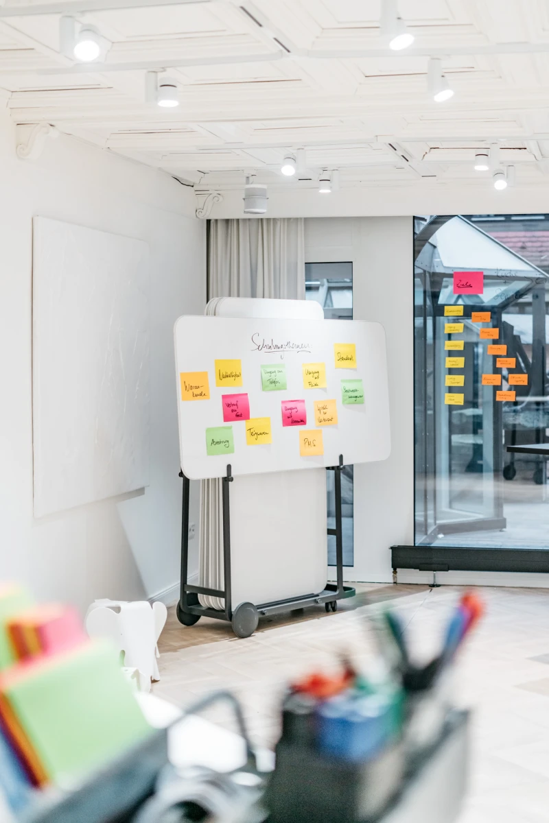 Whiteboard mit bunten Haftnotizen auf einem mobilen St&auml;nder in einem hellen, modernen B&uuml;roraum mit Glaswand und Deckenspots. Verschiedene Farben der Notizen deuten auf eine Brainstorming- oder Planungssitzung hin.