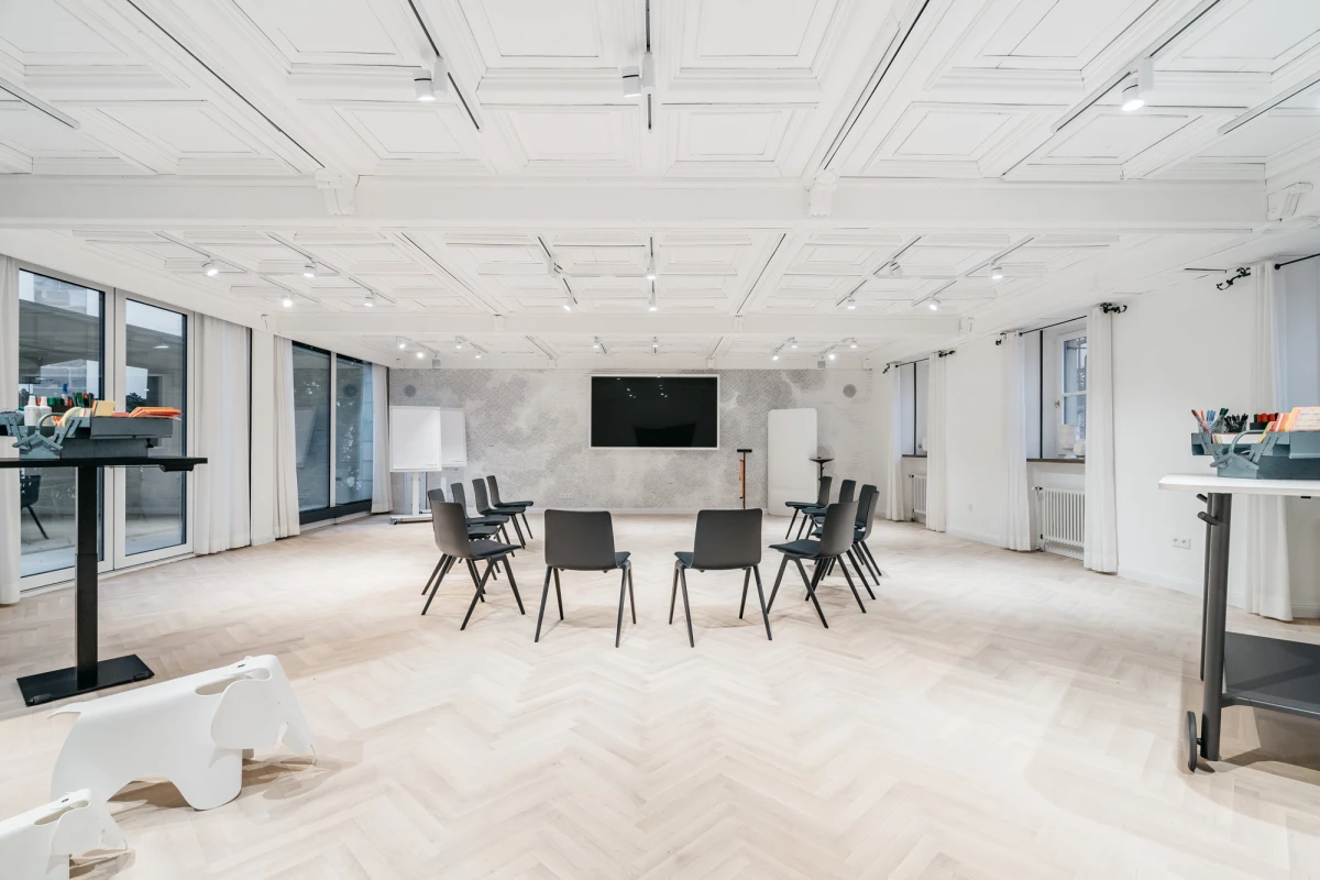 Moderner, heller Schulungsraum mit wei&szlig;en W&auml;nden und Decke. Mehrere schwarze St&uuml;hle sind in einem Halbkreis angeordnet, an der Wand h&auml;ngt ein gro&szlig;er schwarzer Bildschirm. Gro&szlig;e Fenster lassen viel Tageslicht herein und verleihen dem Raum eine offene, einladende Atmosph&auml;re.