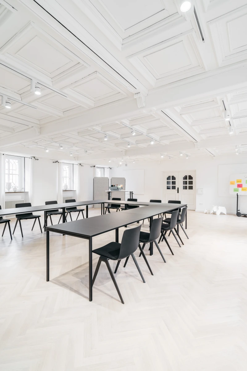 Heller, minimalistischer Schulungsraum mit gro&szlig;en Tischen und schwarzen St&uuml;hlen. Wei&szlig;e Decke mit Holzbalken und gro&szlig;e Fenster sorgen f&uuml;r eine offene, moderne Atmosph&auml;re. Die Einrichtung wirkt schlicht und funktional, ideal f&uuml;r Meetings oder Seminare.
