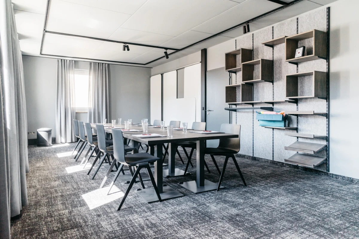 Moderner Konferenzraum mit gro&szlig;em schwarzem Tisch, grauen St&uuml;hlen und strukturierter Teppichoberfl&auml;che. An der Wand offene Regale und Wandschr&auml;nke in gedeckten Farben, gro&szlig;e Fenster mit hellen Vorh&auml;ngen lassen nat&uuml;rliches Licht herein.