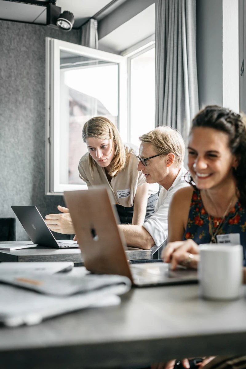 Drei Personen arbeiten konzentriert an Laptops an einem Tisch nahe einem Fenster. Die Stimmung ist entspannt und produktiv, mit einer Kaffeetasse im Vordergrund. Sie scheinen in einem modernen B&uuml;ro oder Besprechungsraum zu sein.