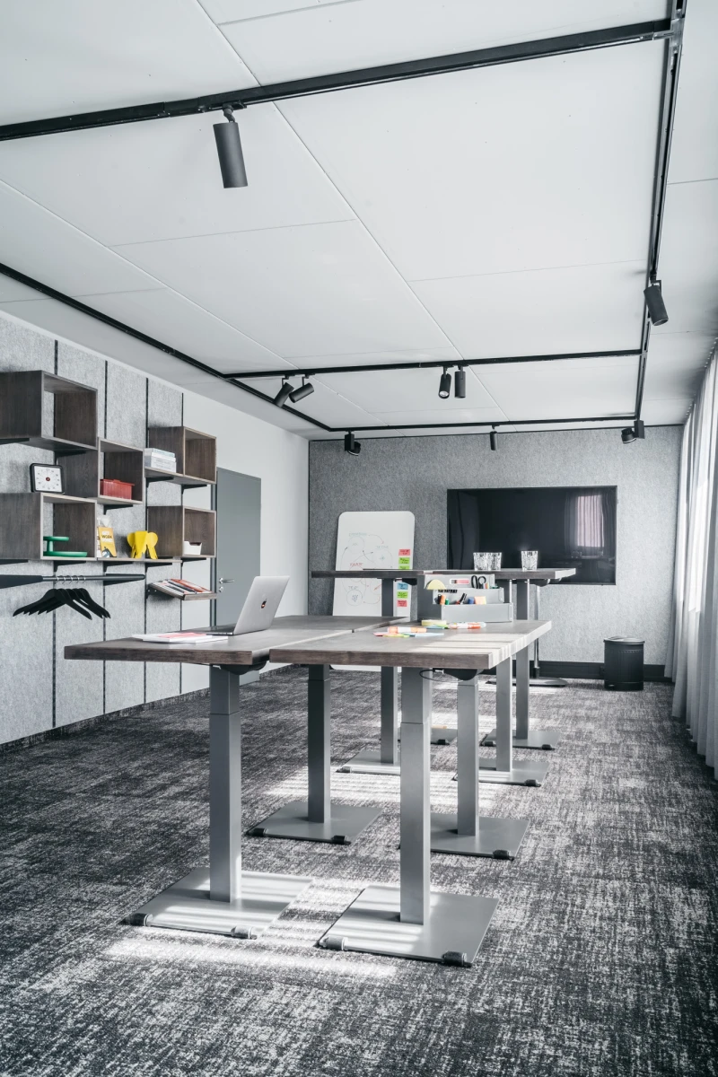 Moderner B&uuml;roraum mit h&ouml;henverstellbaren Schreibtischen, grauen Bodenfliesen und wei&szlig;en Wandregalen. Schwarze Deckenspots erhellen den minimalistischen Arbeitsbereich mit offenem, hellem Raumkonzept.