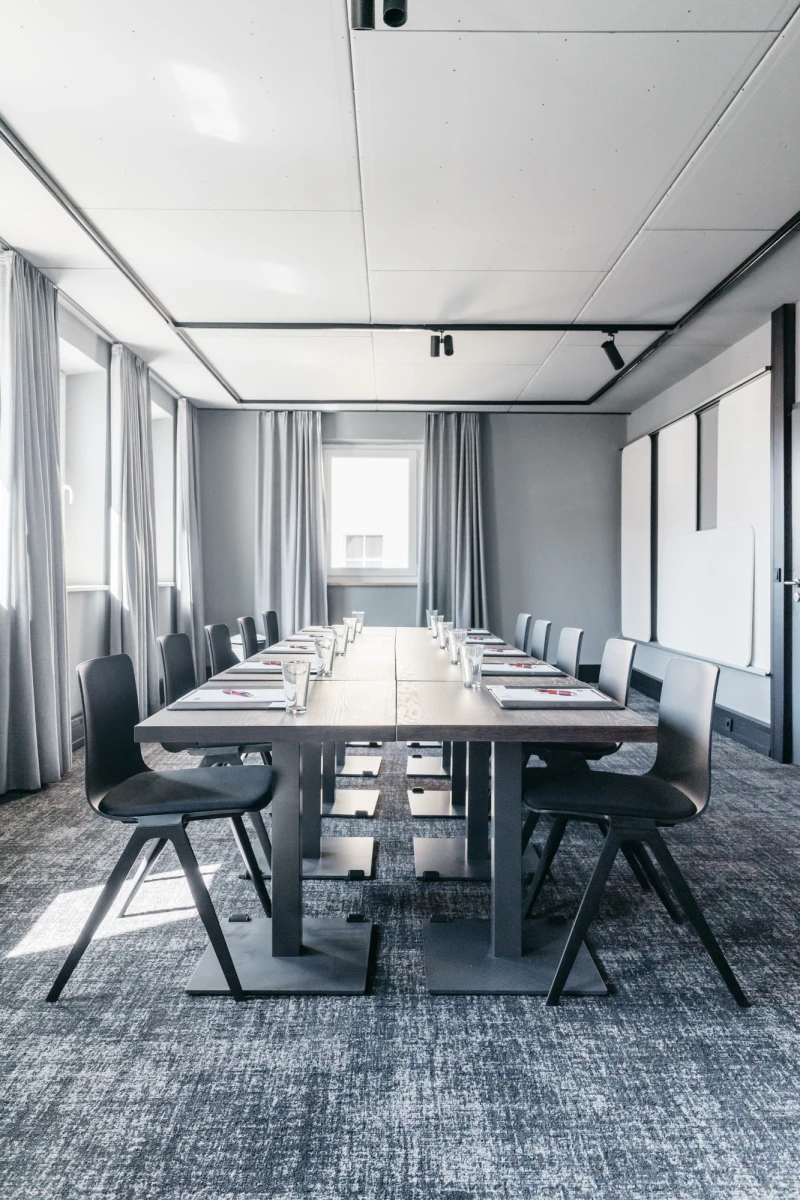 Moderner Konferenzraum mit langem Holztisch und grauen B&uuml;rost&uuml;hlen, umgeben von wei&szlig;en W&auml;nden und bodenlangen Vorh&auml;ngen. Tageslicht f&auml;llt durch die Fenster und erhellt den minimalistischen, professionellen Raum.