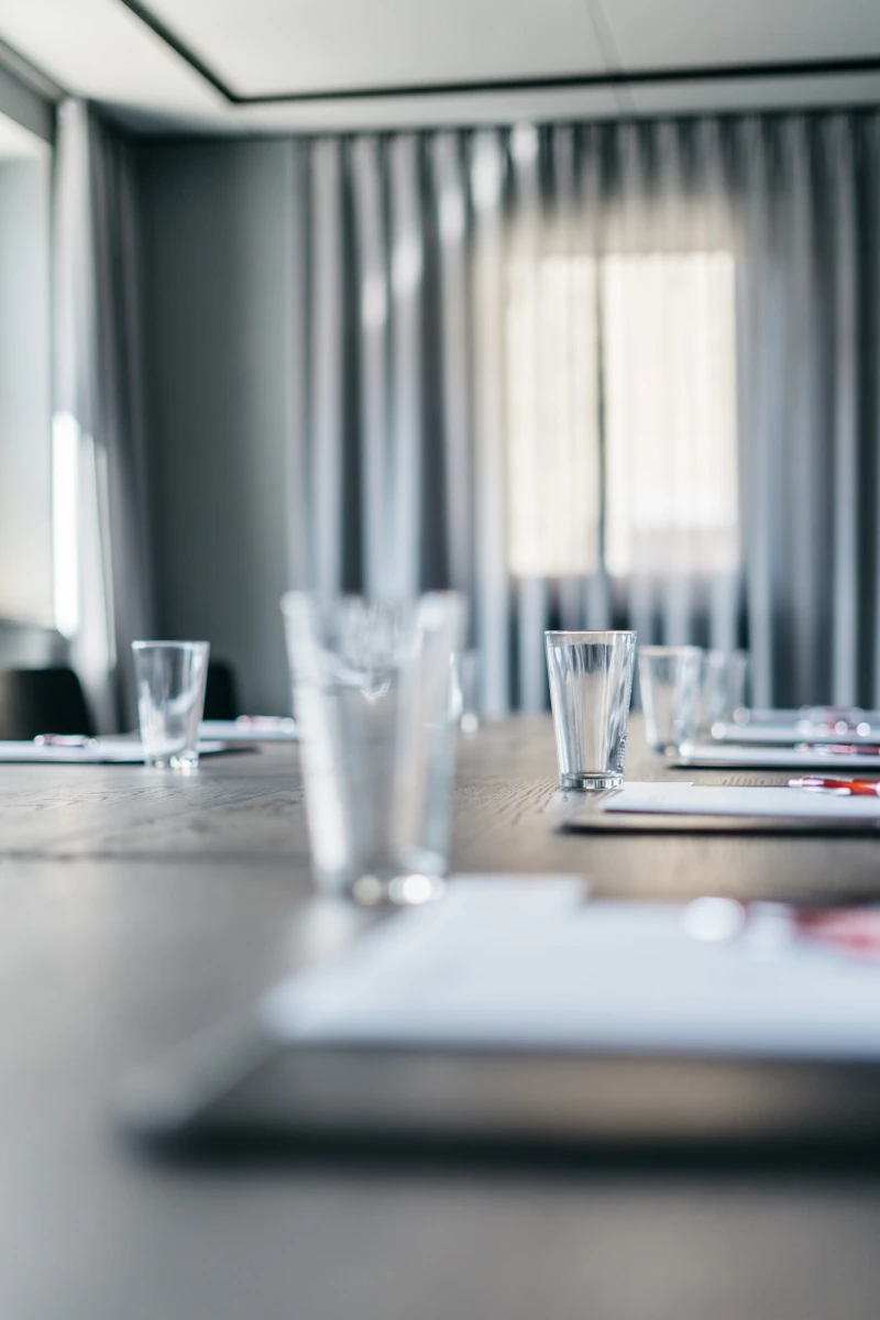 Konferenztisch mit Wassergl&auml;sern und Dokumenten, umgeben von ged&auml;mpften grauen Vorh&auml;ngen und einem unscharfen Fenster im Hintergrund. Professionelle Meetingatmosph&auml;re in einem ruhigen, minimalistischen Besprechungsraum.