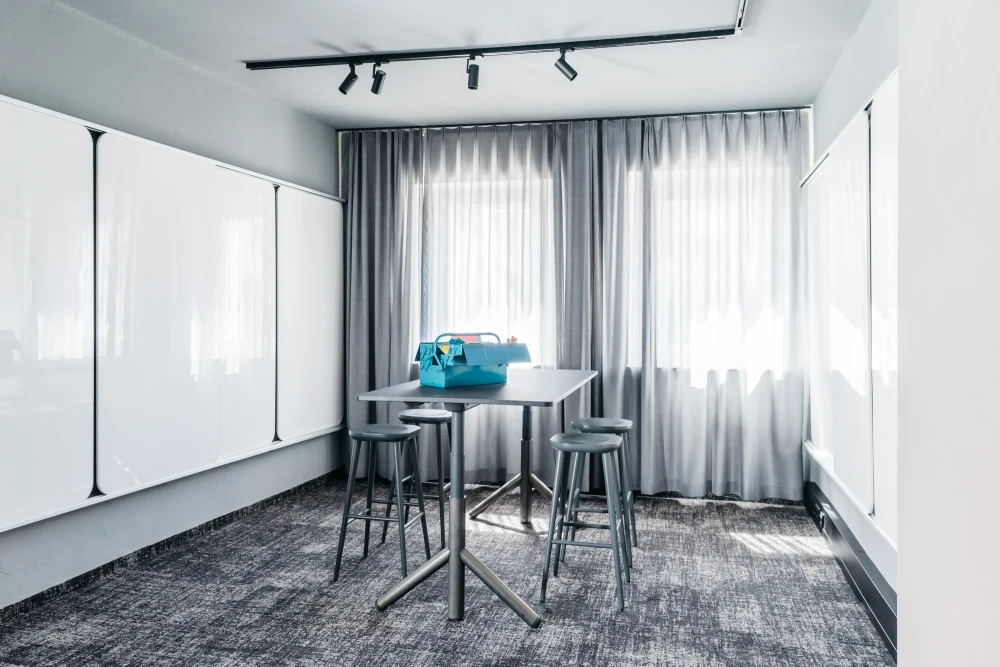 Moderner Konferenzraum mit grauen Vorh&auml;ngen und Teppichboden. Hochstehtische mit Barhockern und eine blaue Tasche stehen bereit. Schwarze Schienenstrahler an der Decke sorgen f&uuml;r optimale Beleuchtung.