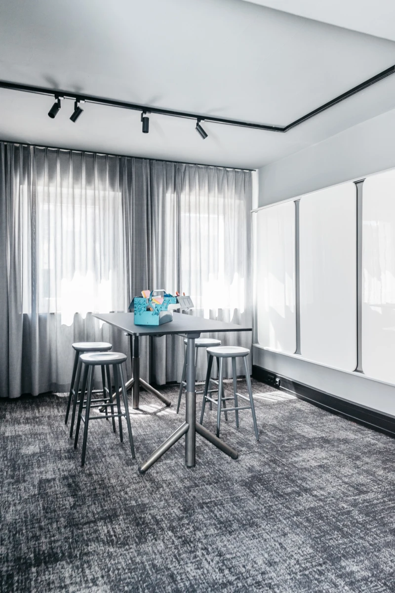 Kleiner Meetingraum mit grauem Teppichboden, einem runden Stehtisch und drei Barhockern. Gro&szlig;e Fenster mit weichen, hellen Vorh&auml;ngen und Deckenspots sorgen f&uuml;r eine helle, moderne Atmosph&auml;re.