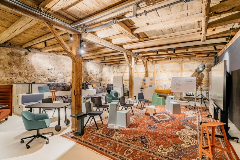 Rustikaler Konferenzraum mit Holzbalkendecke und -st&auml;ndern, ausgestattet mit modernen B&uuml;rost&uuml;hlen und einem gro&szlig;en orangefarbenen Vintage-Teppich. Industrieller Charme trifft auf zeitgen&ouml;ssisches Design in einem umgebauten Scheunenloft mit Steinw&auml;nden und hellen M&ouml;beln.