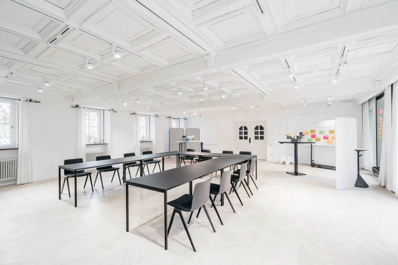 Heller, minimalistischer Seminarraum mit langen schwarzen Tischen und passenden St&uuml;hlen. Wei&szlig;e W&auml;nde und Decke mit Oberlicht, gro&szlig;e Fenster sorgen f&uuml;r nat&uuml;rliches Licht. An der Wand h&auml;ngen bukte Haftnotizen, ein Whiteboard steht bereit f&uuml;r Besprechungen und Workshops.