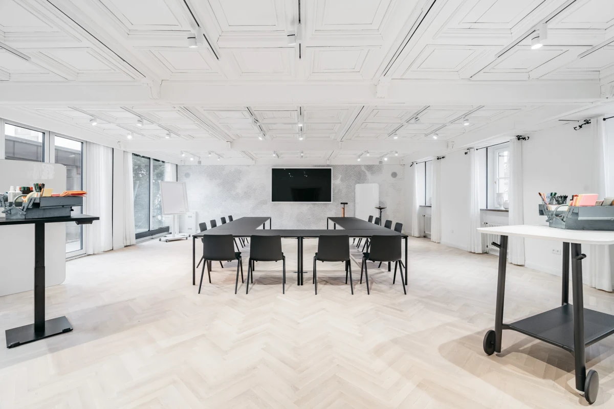 Moderner, heller Konferenzraum mit gro&szlig;er schwarzer Konferenztafel und mehreren schwarzen St&uuml;hlen um einen Tisch. Wei&szlig;e W&auml;nde, gro&szlig;e Fenster und ein heller Holzboden pr&auml;gen den minimalistischen, professionellen Meetingraum.