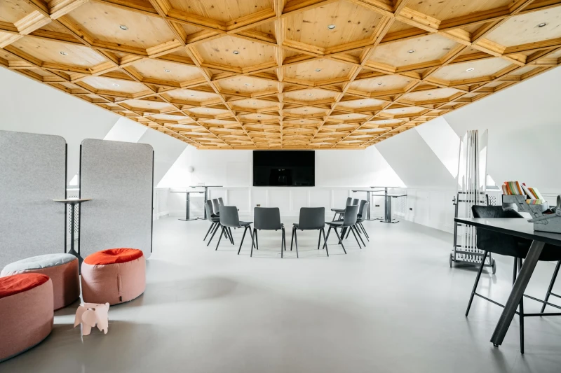 Moderner Konferenzraum mit geometrischer Holzdecke, grauen St&uuml;hlen um einen Tisch und roten Polstersesseln. Ein schwarzer Bildschirm h&auml;ngt an der Wand, und der Raum hat eine helle, minimalistisch gestaltete Atmosph&auml;re.