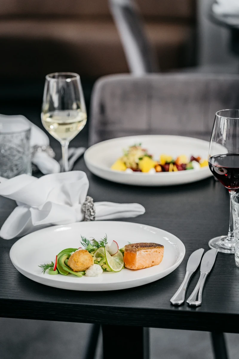 Elegante Restaurantteller mit zartem Lachsfilet, begleitet von einem Wei&szlig;weinglas und einem Rotwein. Fein angerichtet auf einem dunklen Tisch mit wei&szlig;er Serviette und Besteck, eine geschmackvolle Komposition aus Gourmet-K&uuml;che.