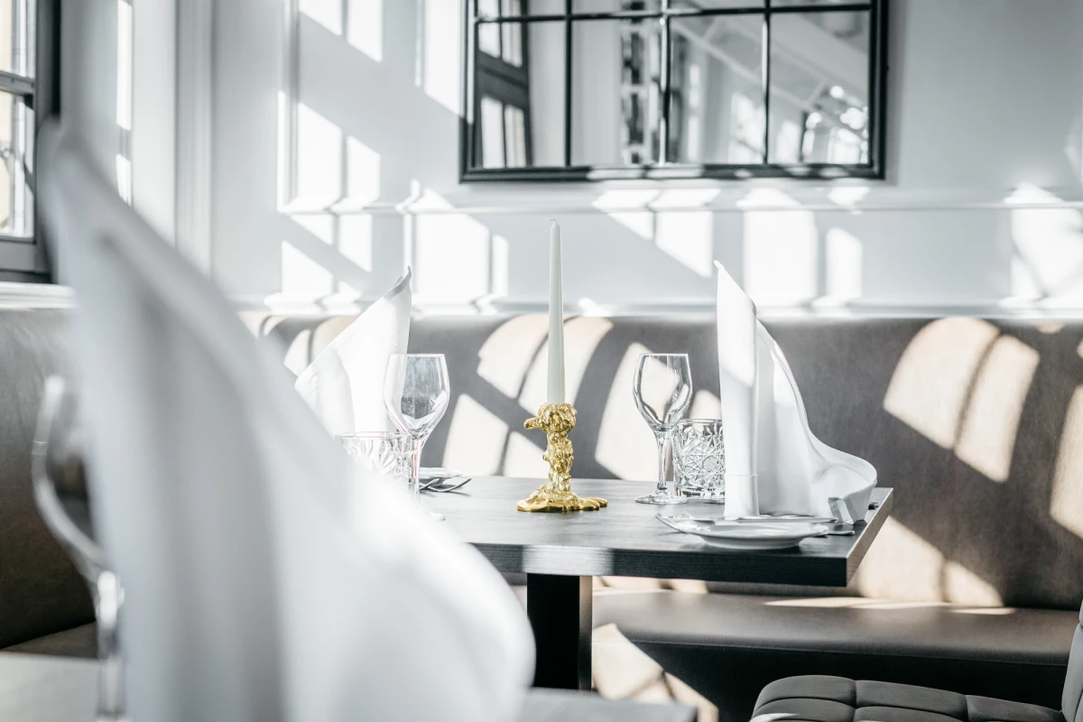 Elegante Tischdekoration in einem hellen, modernen Restaurant-Ambiente mit gedeckten Tischen, wei&szlig;en St&uuml;hlen und gro&szlig;en Fenstern, die weiches Tageslicht hereinlassen.