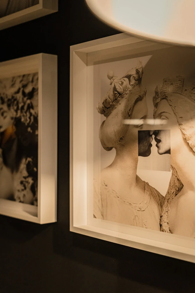 Vintage Schwarzwei&szlig;fotografie in einem wei&szlig;en Rahmen, die eine z&auml;rtliche Umarmung und einen Kuss zweier Silhouetten zeigt, aufgeh&auml;ngt an einer dunklen Wand neben einem weiteren gerahmten Bild.