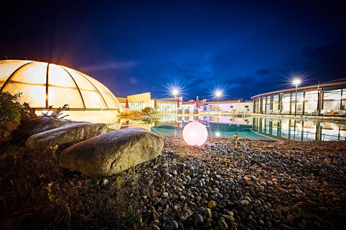 N&auml;chtliche Ansicht der Frankentherme mit beleuchtetem Au&szlig;enbecken, Kieselsteinen im Vordergrund und einer gl&uuml;henden Kugel, umgeben von modernen Geb&auml;uden mit Lichtern und blauem Abendhimmel.