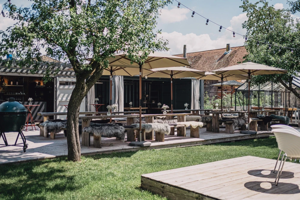 Sonnige Gartenterrasse eines Landhotels mit Sonnenschirmen, Holzm&ouml;beln und einem gro&szlig;en Baum, die zum Verweilen einl&auml;dt. Gepflegter Rasenbereich und rustikale Geb&auml;ude im Hintergrund schaffen eine einladende Atmosph&auml;re.