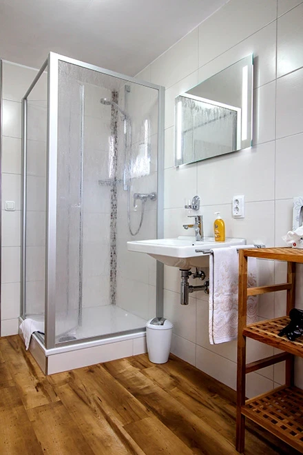 Modernes, helles Badezimmer mit Duschkabine aus Glas, wei&szlig;en Fliesen und Holzboden. Holzregal mit Handtuch, wei&szlig;es Waschbecken und Spiegel an der Wand, gelbe Duschutensilien auf der Ablage.