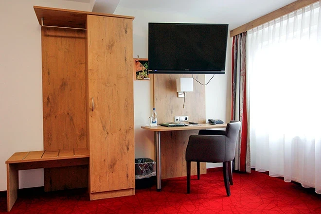 Hotelzimmer mit rotem Teppich, Holzschrank, Schreibtisch mit schwarzem Stuhl und an der Wand montiertem Flach-TV. Helle Gardinen lassen nat&uuml;rliches Licht herein, und die Einrichtung wirkt funktional und modern.