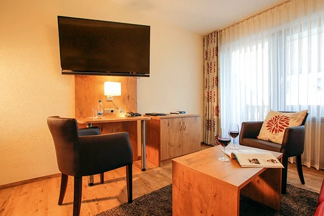 Hotelzimmer mit Holzm&ouml;beln, Fernseher an der Wand und einem Schreibtisch mit B&uuml;rostuhl. Ein Sessel mit gemustertem Kissen steht neben einem Couchtisch mit Weinglas und Zeitschrift, gro&szlig;e Fenster mit hellen Vorh&auml;ngen lassen nat&uuml;rliches Licht herein.