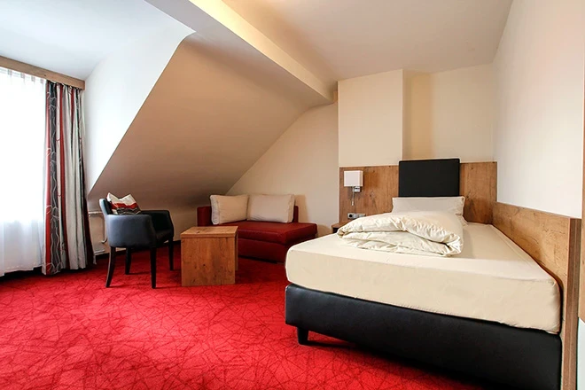 Hotelzimmer mit rotem Teppich, schr&auml;ger Decke und einem Einzelbett. Holzm&ouml;bel und ein blauer Sessel komplettieren die gem&uuml;tliche Einrichtung. Das Zimmer ist hell und freundlich gestaltet.