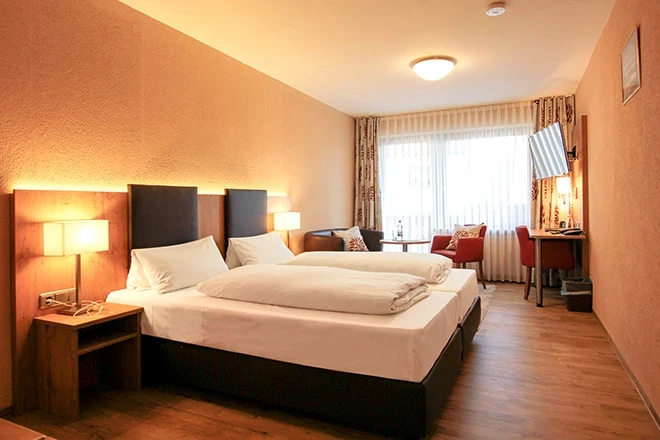 Hotelzimmer mit gro&szlig;em Bett, wei&szlig;er Bettw&auml;sche und gepolstertem Kopfteil. Warme, orangefarbene Beleuchtung und Vorh&auml;nge, daneben ein roter Sessel und Holzm&ouml;bel, helle Fensterfront mit Tageslicht.