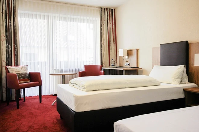 Modernes Hotelzimmer mit einem gro&szlig;en Bett, zwei roten Sesseln und bodenlangen Vorh&auml;ngen vor einem hellen Fenster. Der Raum hat einen roten Teppich und einen dunklen Holzboden, und das Bett ist mit wei&szlig;er Bettw&auml;sche bezogen.