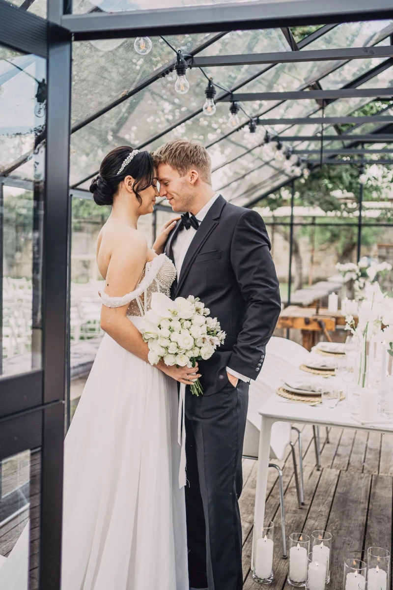 Braut und Br&auml;utigam stehen in einem lichtdurchfluteten Glashaus, umgeben von eleganter Hochzeitsdekoration. Sie tr&auml;gt ein langes wei&szlig;es Kleid und h&auml;lt einen wei&szlig;en Blumenstrauss, w&auml;hrend er einen schwarzen Anzug mit Fliege tr&auml;gt.