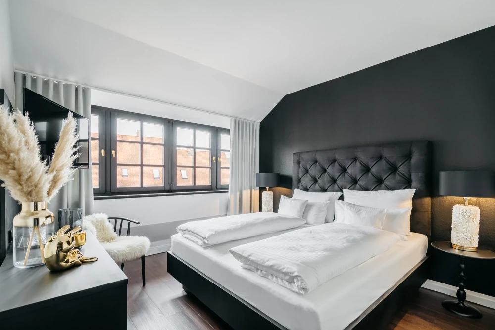 Modernes Hotelzimmer mit grauer Akzentwand und gepolstertem Kopfteil. Gro&szlig;es Bett mit wei&szlig;er Bettw&auml;sche, Nachttischlampen und Pampasgrasdekoration am Fenster. Helle Atmosph&auml;re mit Holzboden und gro&szlig;em Fenster mit Vorh&auml;ngen.