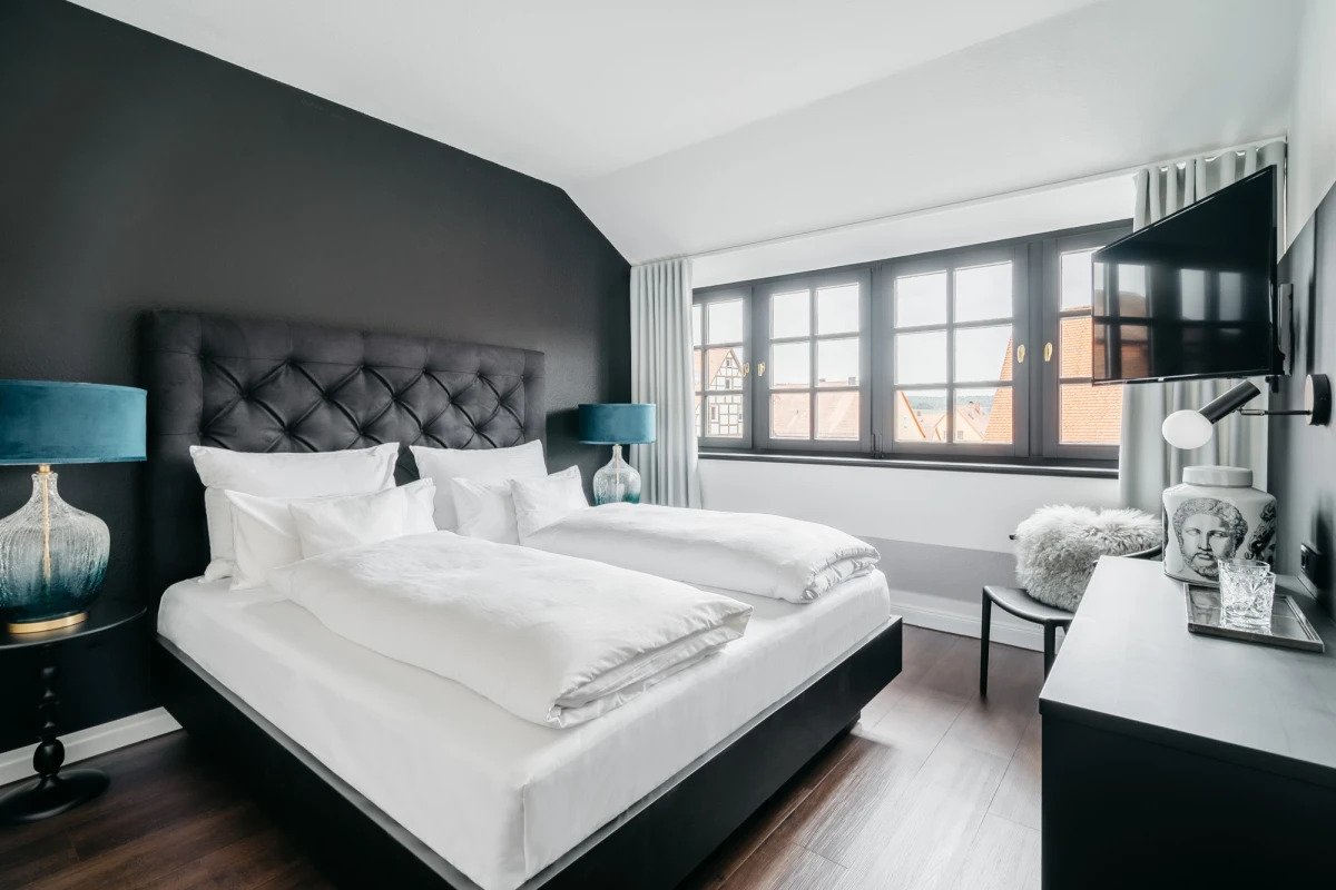Elegant eingerichtetes Hotelzimmer mit schwarzer Akzentwand, gepolstertem Kopfteil und wei&szlig;er Bettw&auml;sche. Gro&szlig;e Fensterfront l&auml;sst nat&uuml;rliches Licht herein, neben dem Bett stehen blaue Designerlampen auf Nachttischen. Modernes Interieur mit dunklem Holzboden und TV an der Wand.