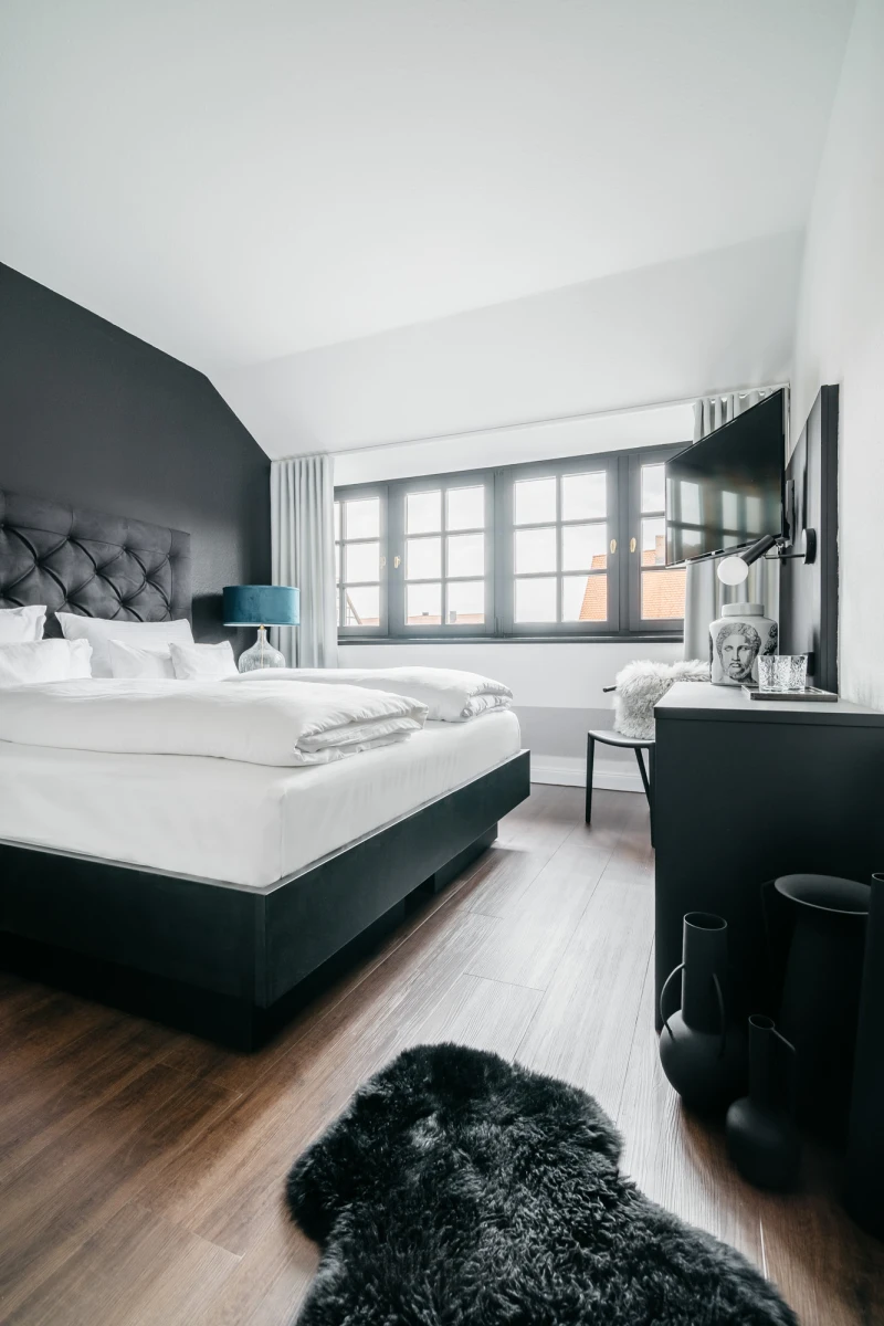 Ger&auml;umiges Hotelzimmer mit gro&szlig;em gepolstertem Bett in Anthrazit, hellen Bettw&auml;schekissen und Holzboden. Gro&szlig;es Fenster mit Vorh&auml;ngen, Flachbild-TV an der Wand und ein schwarzer Fellteppich im Vordergrund. Modernes, minimalistisches Designambiente in ged&auml;mpften Graut&ouml;nen.