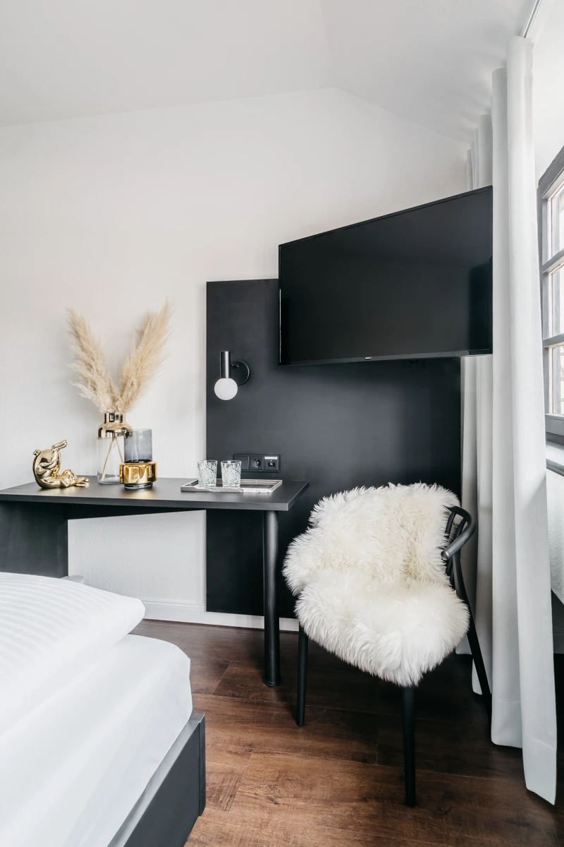 Modernes Hotelzimmer mit schwarzem Schreibtisch und wei&szlig;em Fell auf einem Stuhl. Pampas-Gras in einer Vase und goldene Dekoartikel zieren den Raum, der helle W&auml;nde und einen Holzboden hat.