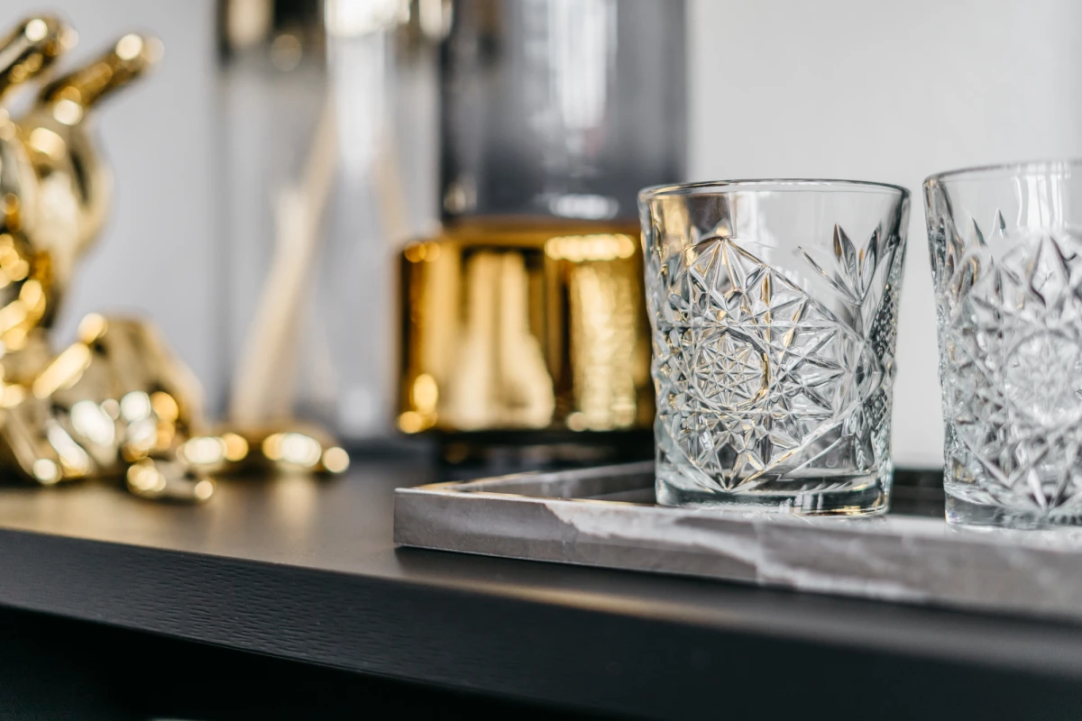 Zwei geschliffene Whiskygl&auml;ser auf einem dunklen Tablett mit verschwommenen goldenen Dekorationselementen im Hintergrund. Die Gl&auml;ser haben ein aufw&auml;ndiges, geometrisches Schliffmuster und stehen bereit f&uuml;r einen eleganten Drink.