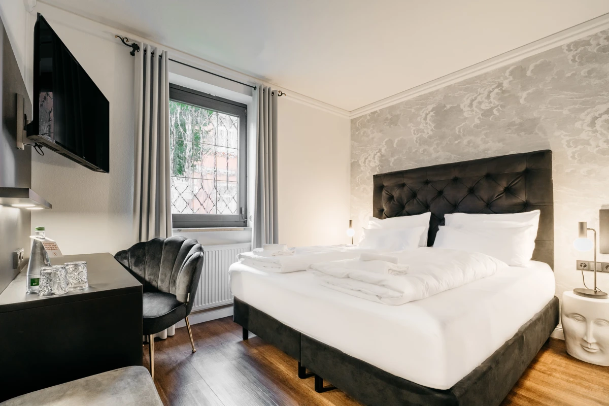 Modernes Hotelzimmer mit gro&szlig;em gepolstertem Bett, dunkelgrauem Kopfteil und wei&szlig;er Bettw&auml;sche. Daneben ein Schreibtisch mit einem grauen Sessel, an der Wand ein Flachbild-Fernseher und ein Fenster mit Gardinen. Der Boden ist aus dunklem Holz und die W&auml;nde sind in neutralen Farbt&ouml;nen gehalten.