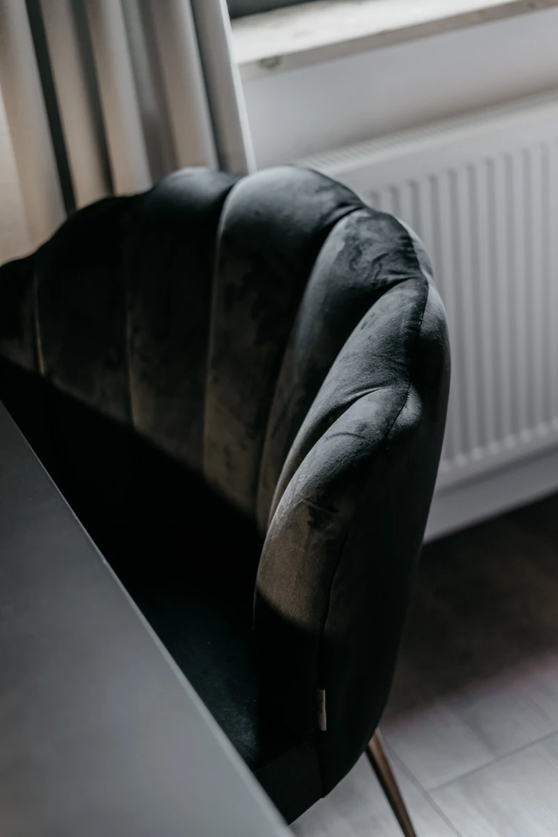 Schwarzer Sessel mit weichen Polstern an der Lehne, der vor einem wei&szlig;en Heizk&ouml;rper und einem Fenster mit Vorhang steht. Der Stuhl hat einen geschwungenen, eleganten Designerstil und wirkt edel und modern.
