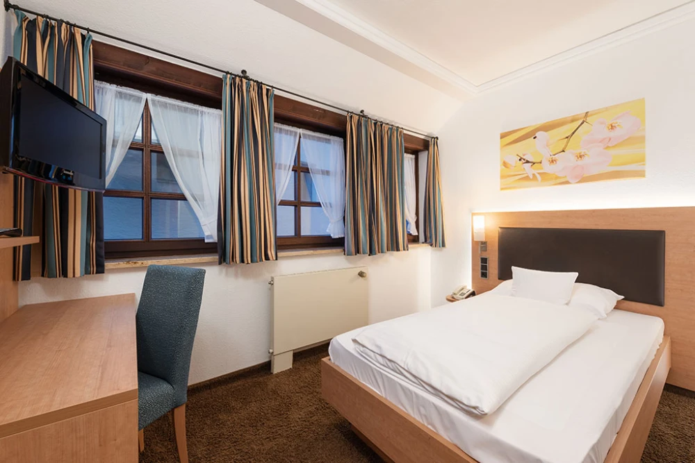 Gem&uuml;tliches Einzelzimmer mit Holzm&ouml;beln, wei&szlig;em Bett, blau-beigen gestreiften Vorh&auml;ngen und gro&szlig;en Fenstern. Auf dem Schreibtisch steht ein Flat-TV, und ein gepolsterter Stuhl steht daneben. An der Wand h&auml;ngt ein gelbes Kunstwerk &uuml;ber dem Bett.