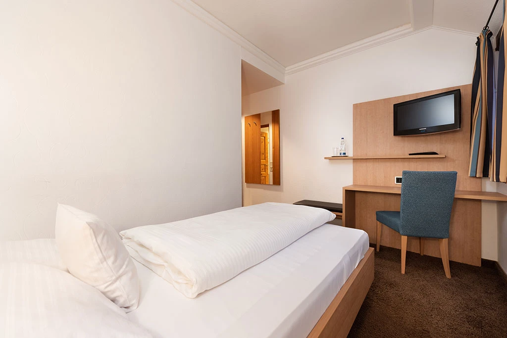 Schlichtes Hotelzimmer mit wei&szlig;em Bett, blauem Stuhl und Flachbild-TV an der Holzwand. Helle, freundliche Atmosph&auml;re mit Holzm&ouml;beln und Zugang zum Badezimmer.