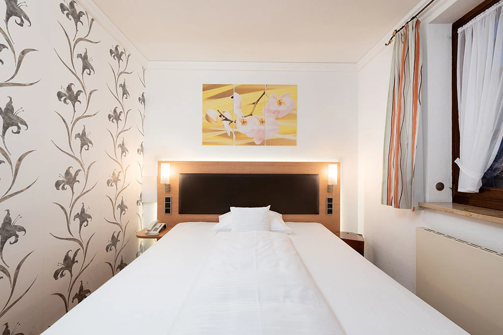 King-Size-Bett in einem hell eingerichteten Hotelzimmer mit gemusterten Tapeten, einem gelben Kunstdruck &uuml;ber dem Bett und gestreiften Vorh&auml;ngen am Fenster. Die wei&szlig;e Bettw&auml;sche liegt ordentlich aufgeschlagen, und die Beleuchtung am Kopfteil sorgt f&uuml;r eine einladende Atmosph&auml;re.