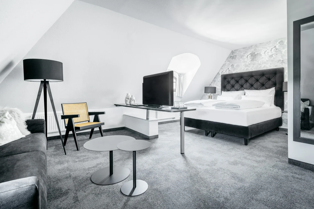 Modernes Hotelzimmer mit gro&szlig;em Doppelbett, gepolstertem Sessel und Stehlampe auf grauem Teppichboden. Wei&szlig;e W&auml;nde und minimalistische Einrichtung in Grau- und Wei&szlig;t&ouml;nen, mit Flachbild-TV und eleganten M&ouml;beln.