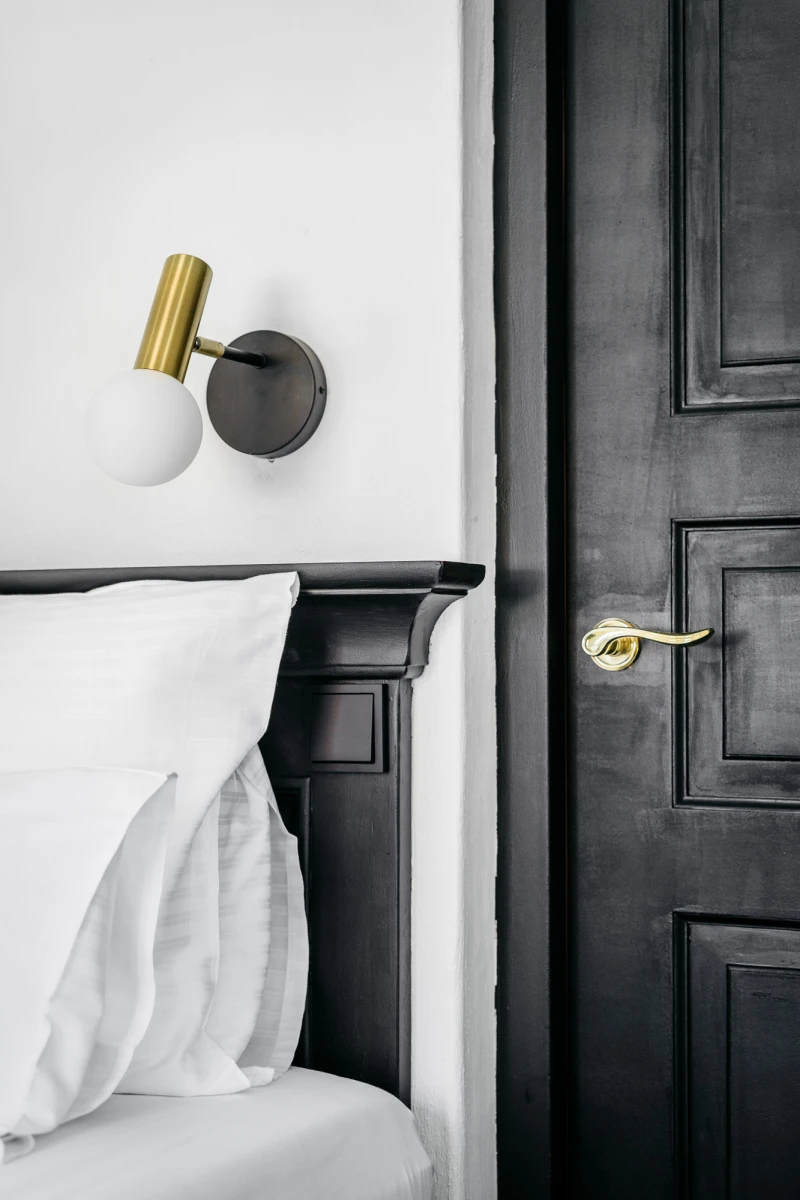 Modernes Hotelzimmer mit elegantem Schlafbereich, schwarzer T&uuml;r mit goldener Klinke und einer goldenen Wandleuchte an der wei&szlig;en Wand, die minimalistisches und stilvolles Interieurdesign betont.