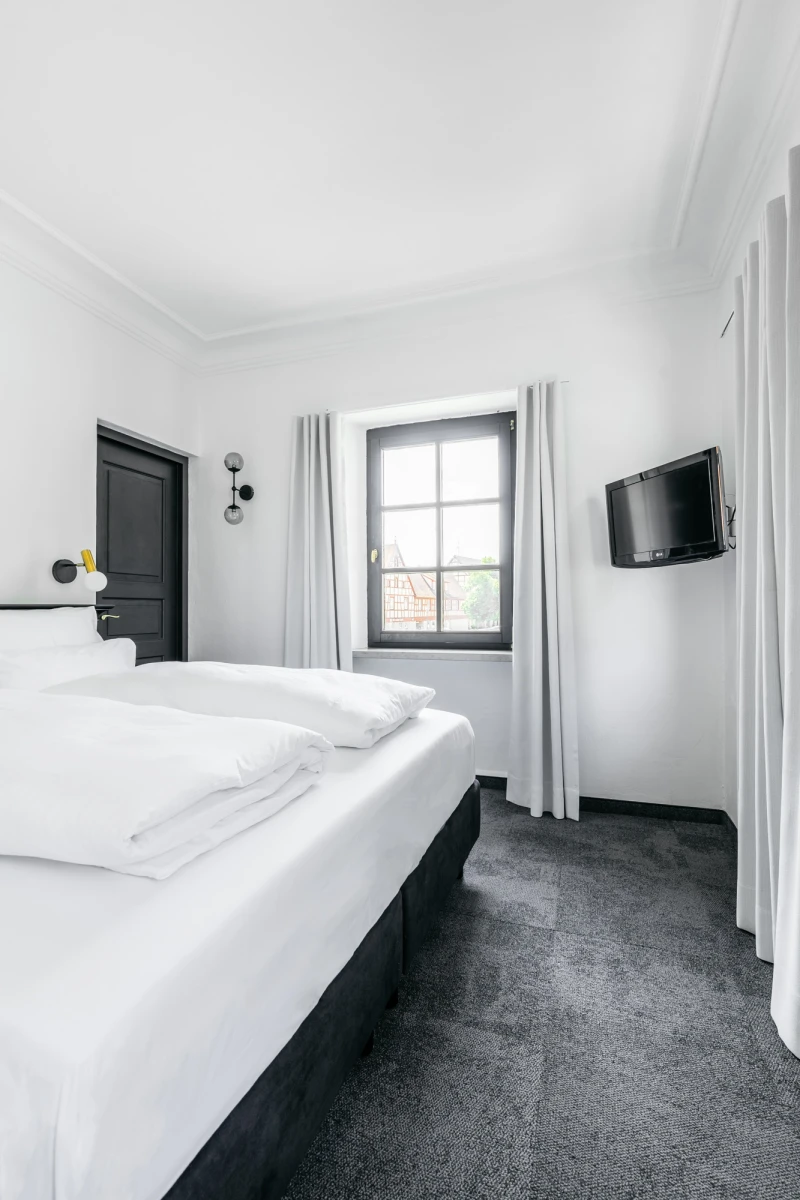 Modernes Hotelzimmer in Grau und Wei&szlig; mit einem gro&szlig;en wei&szlig;en Bett, dunkelgrauem Teppichboden und einer Fensterfront. An der Wand h&auml;ngt ein Flachbild-Fernseher, und durch das Fenster f&auml;llt ged&auml;mpftes Tageslicht.