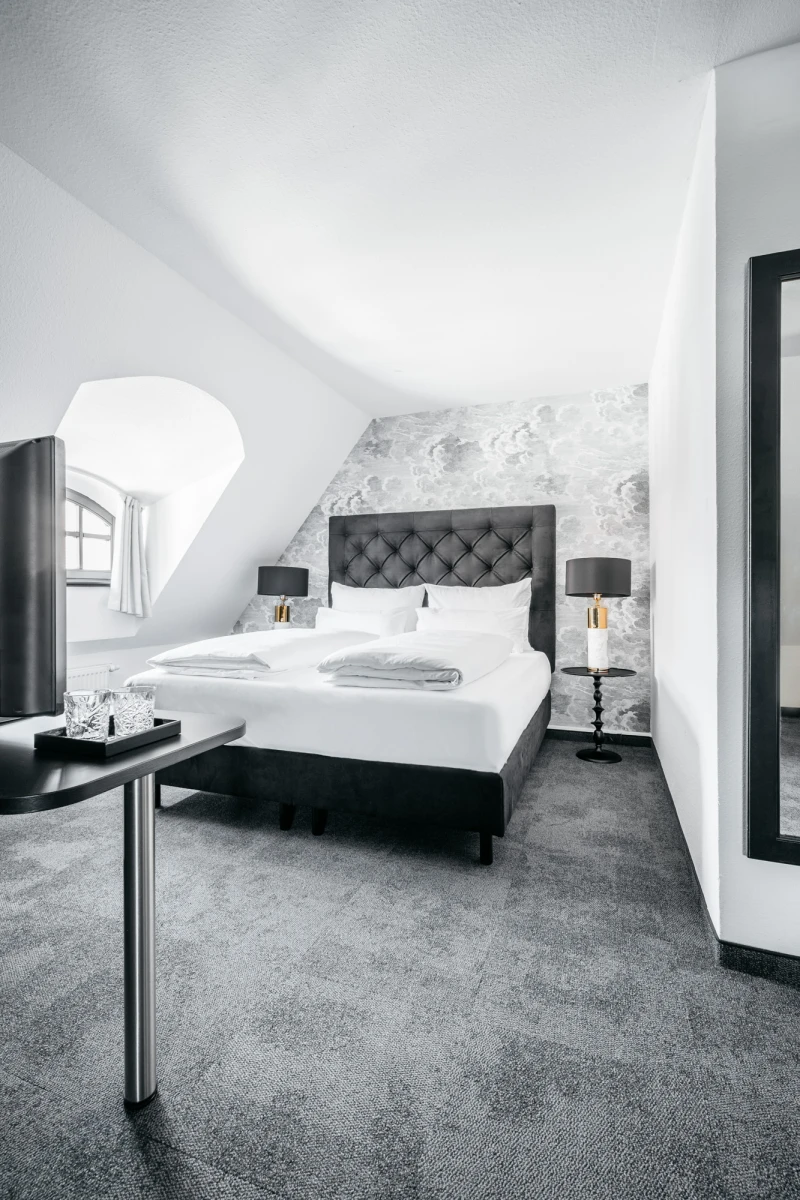 Hotelzimmer im Dachgeschoss mit einem gro&szlig;en grauen Bett, gepolstertem Kopfteil und schwarzen Nachttischen. Graue Teppichb&ouml;den, wei&szlig;e W&auml;nde und ein Dachfenster sorgen f&uuml;r eine helle, moderne Atmosph&auml;re.