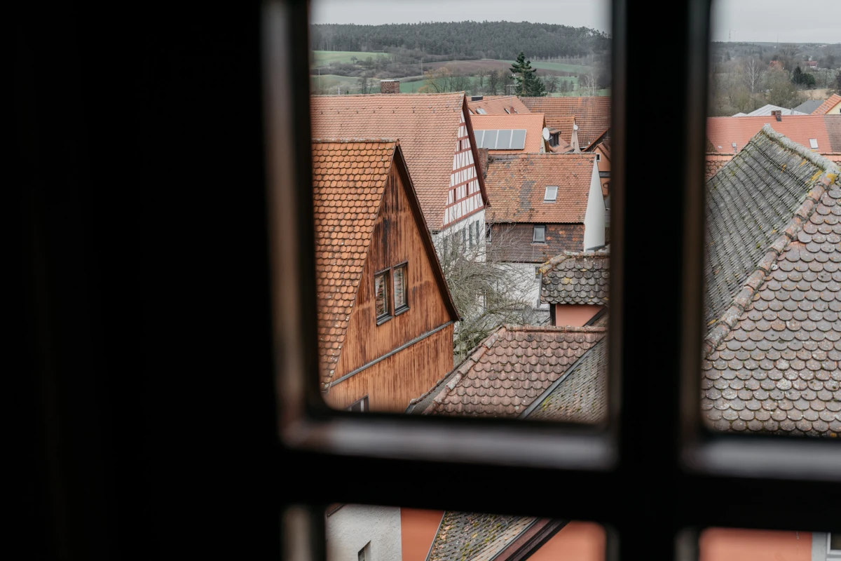 Blick durch ein Fenster auf alte D&auml;cher mit Ziegeln in verschiedenen Braunt&ouml;nen, charakteristisch f&uuml;r eine historische Altstadt mit mittelalterlicher Architektur. Im Hintergrund sanfte H&uuml;gellandschaft und bewachsene Fl&auml;chen.