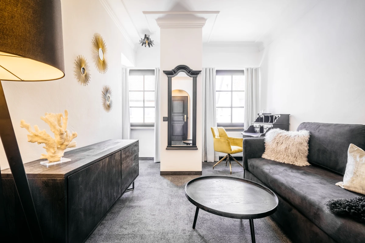 Modernes, helles Hotelzimmer mit grauem Sofa, gelbem Sessel und dekorativen Wandelementen. Wei&szlig;e W&auml;nde, gro&szlig;e Fenster und ein runder Couchtisch pr&auml;gen den minimalistischen, einladenden Wohnbereich. Eine stehende Lampe und eine Kommode vervollst&auml;ndigen das zeitgen&ouml;ssische Interieur.