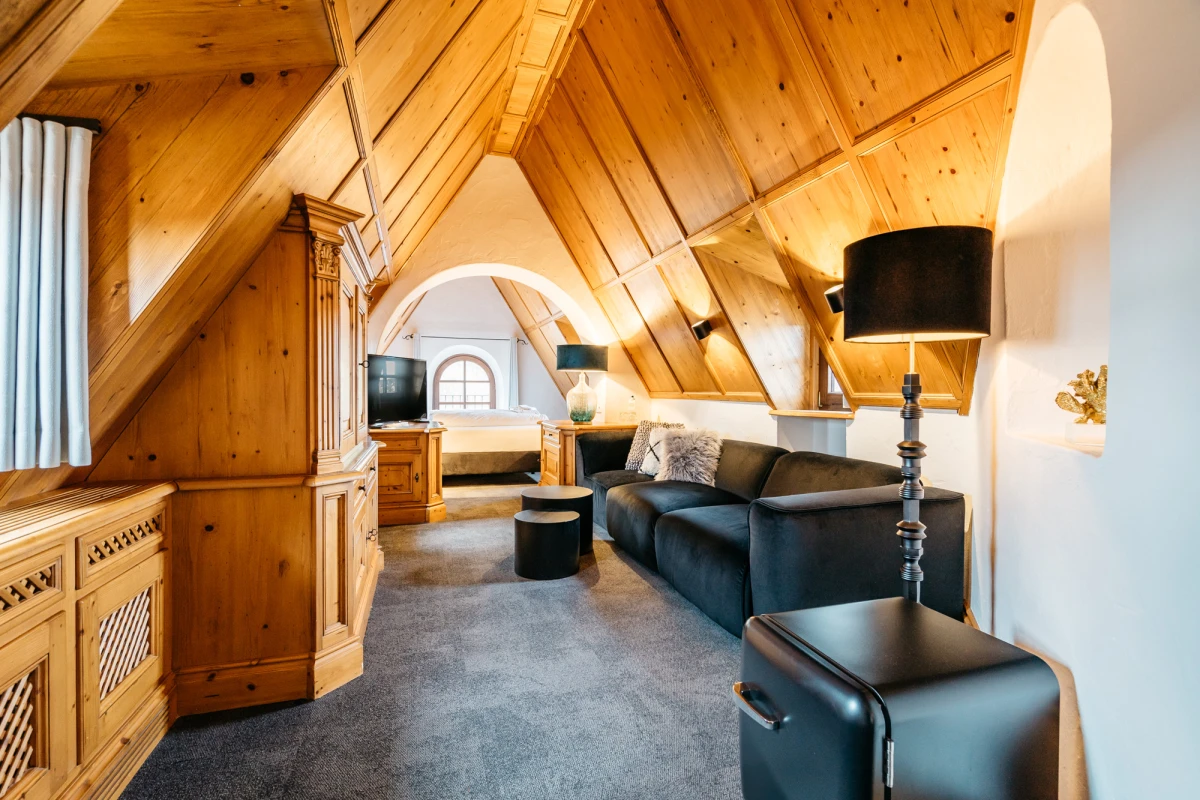 Moderner Dachboden-Wohnbereich mit warmen Holzw&auml;nden und -decke, ausgestattet mit dunkelgrauer Ledercouch, schwarzen Beistelltischen und einer Stehlampe. Der Raum wirkt gem&uuml;tlich und hell durch das Dachfenster und die nat&uuml;rliche Holzoptik.